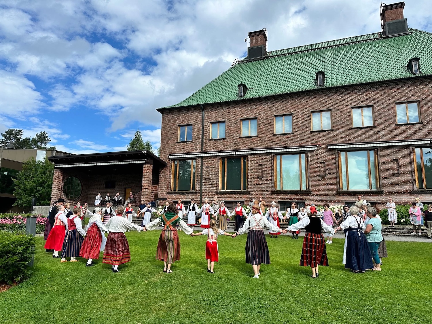 Kansallispukuiset naiset tanssivat piirissä. Taustalla näkyy vanha kartanorakennus.