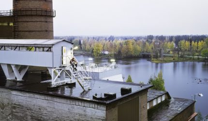 Photograph of Ville Lenkkeri depicting autumn landscape in Mänttä by the name Morning Coffee on a Roof of a Town. A man is sitting on metal staircase on the roof of a paper mill enjoying his newspaper and coffee.