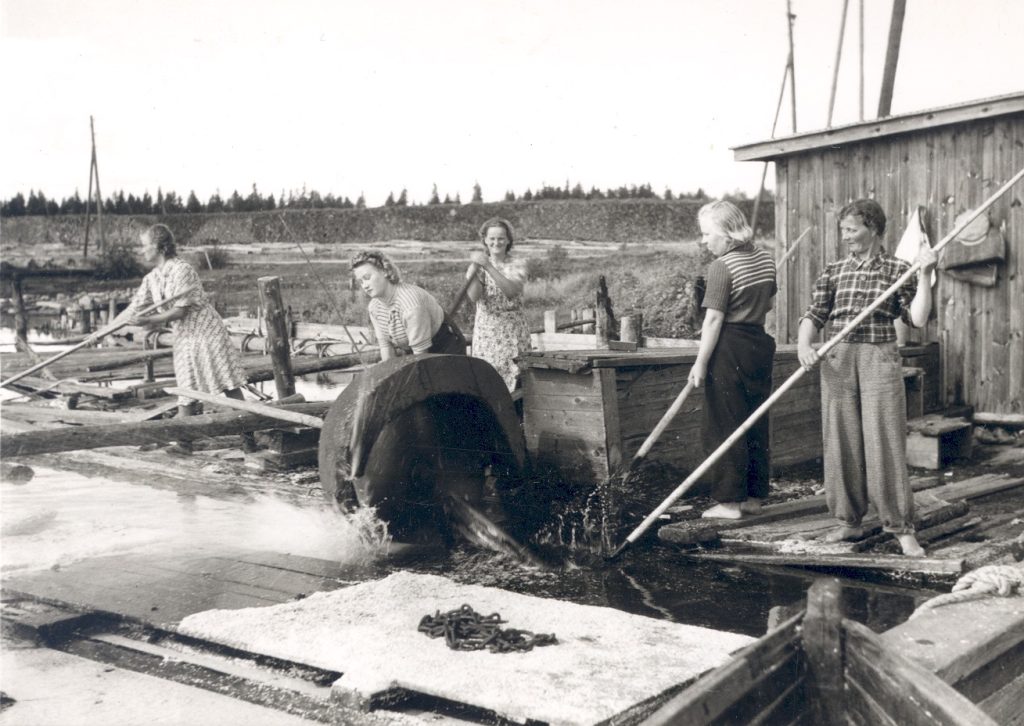 Old black-and-white photograph with smiling women standing at a circular saw on lakeshore with pike poles in their hands.