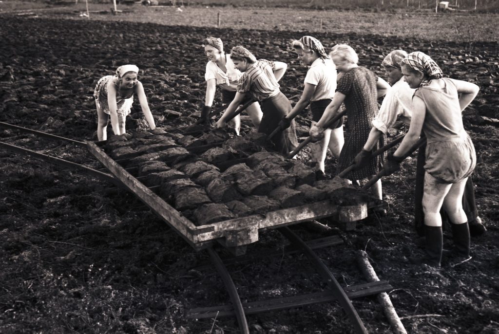 Old black-and-white photograph depicting women loading peat on a railway carriage with skirts and trouser rolled up and wearing scarfs on their head.