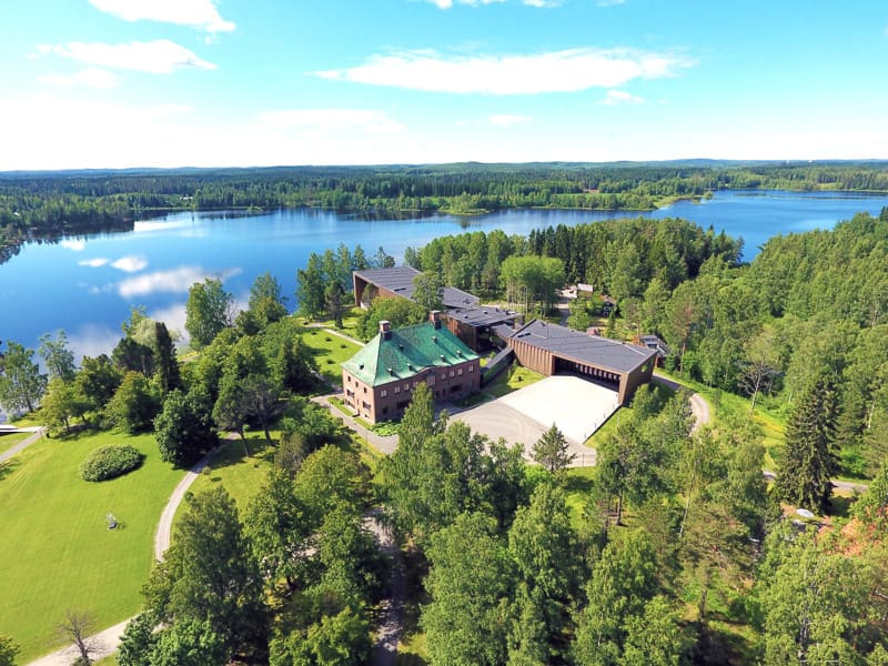 Ilmakuva puiden ympäröivästä suuresta talosta, jonka taustalla on järvi.