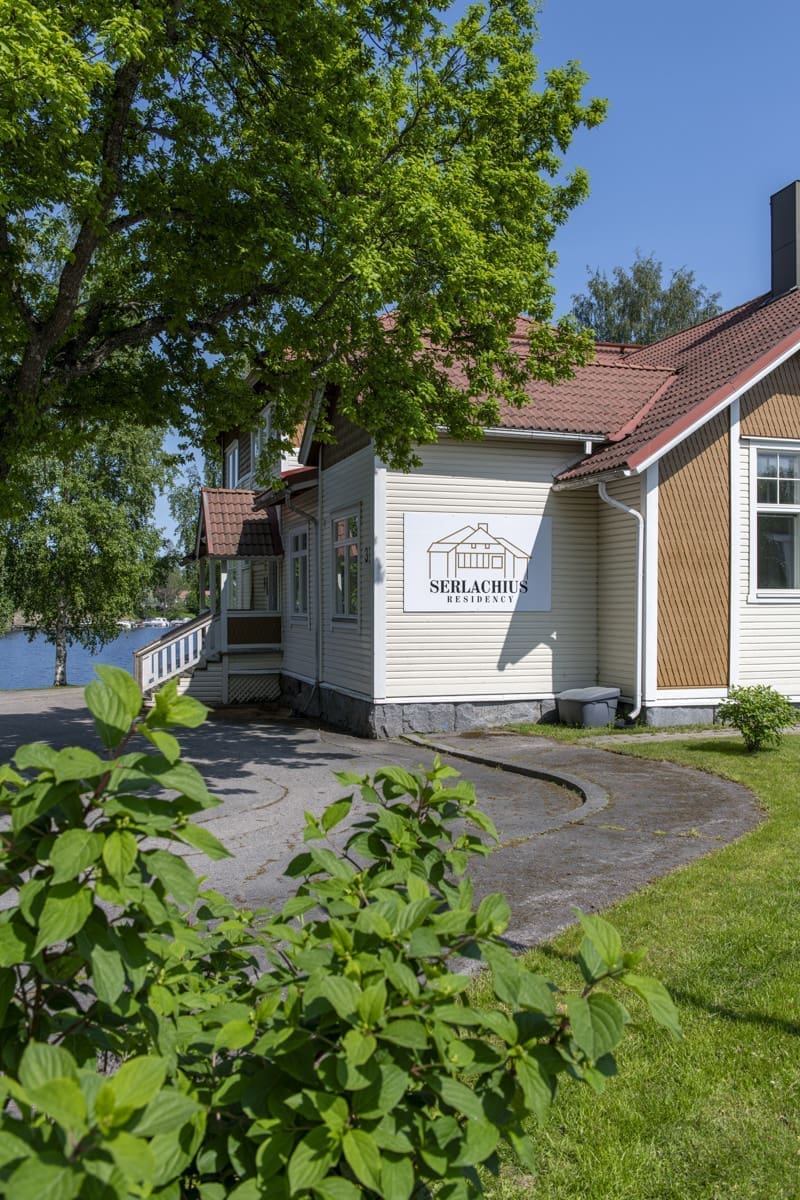 Puurakennus, jonka seinässä on Serlachius Residenssin kyltti, Etualalla vehreitä kasveja.