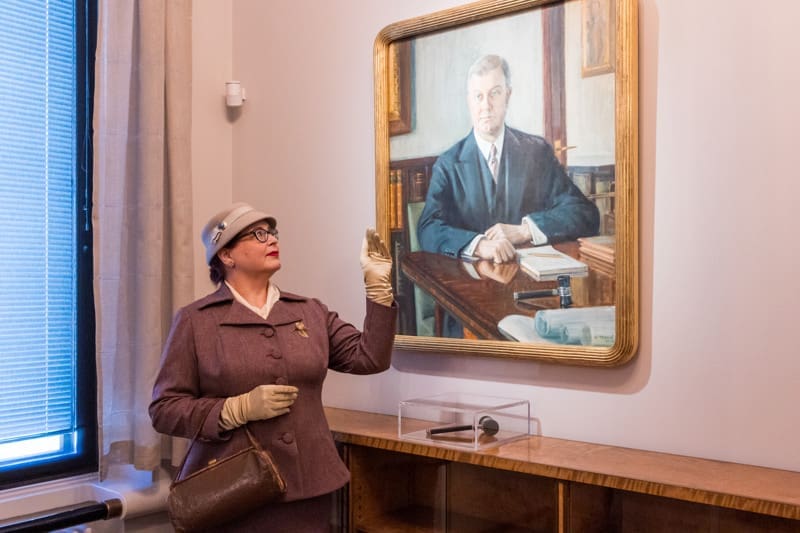 Vanhaan, tyylikkäseen asuun pukeutunut nainen osoittaa muotokuvamaalausta historiallisessa kokoushuoneessa. 