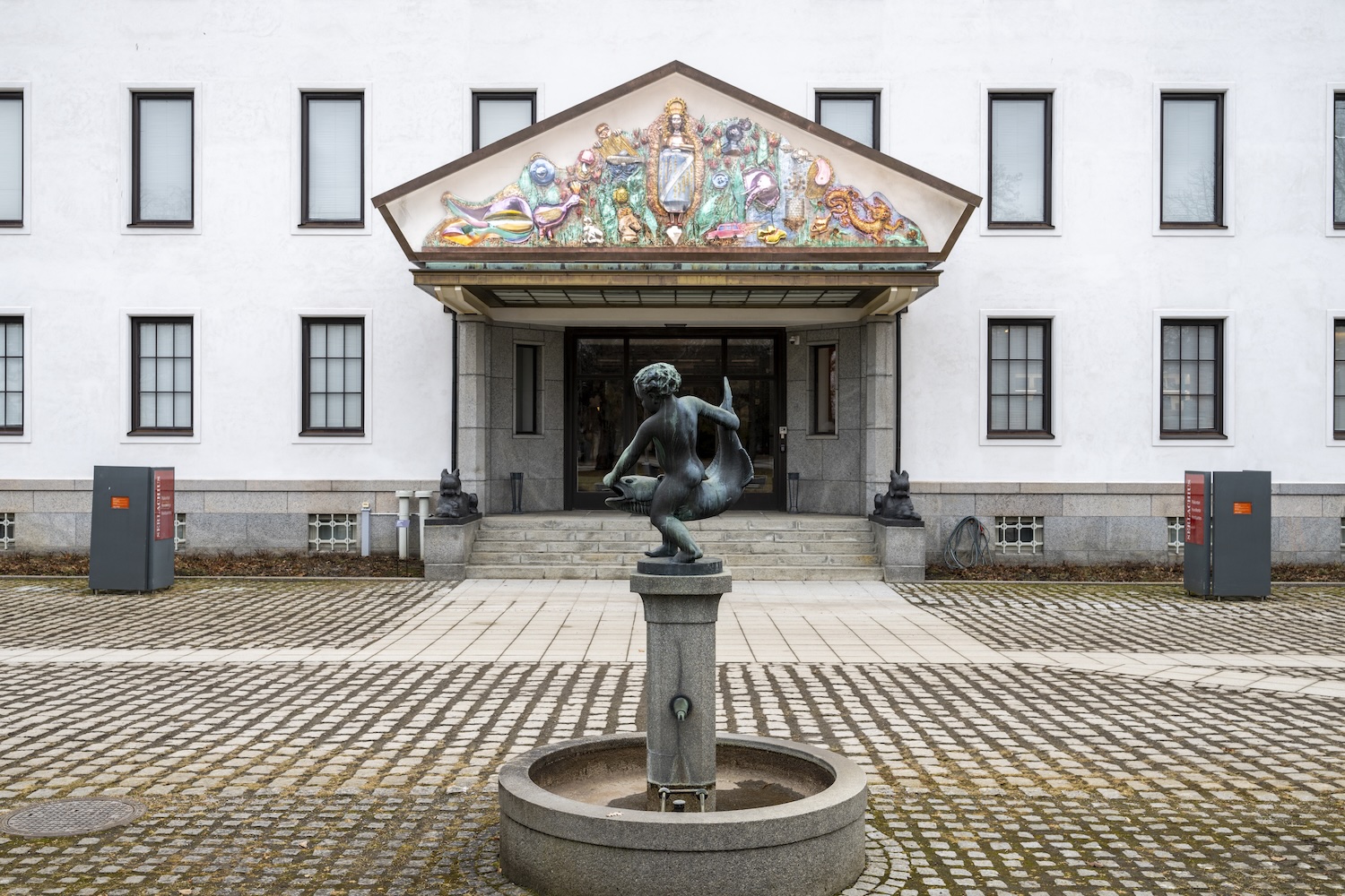 Koristeellinen pahvireliefi valkean rakennuksen pääoven yläpuolella.