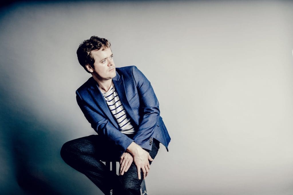 Benjamin Grosvenor istuu korkealla jakkaralla ja katsoo viistoon.