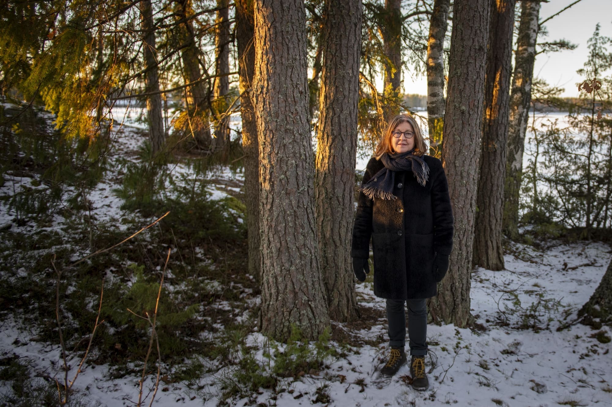 Taiteilija Eija-Liisa Ahtila seisoo talvisessa metsässä, johon paistaa aurinko.