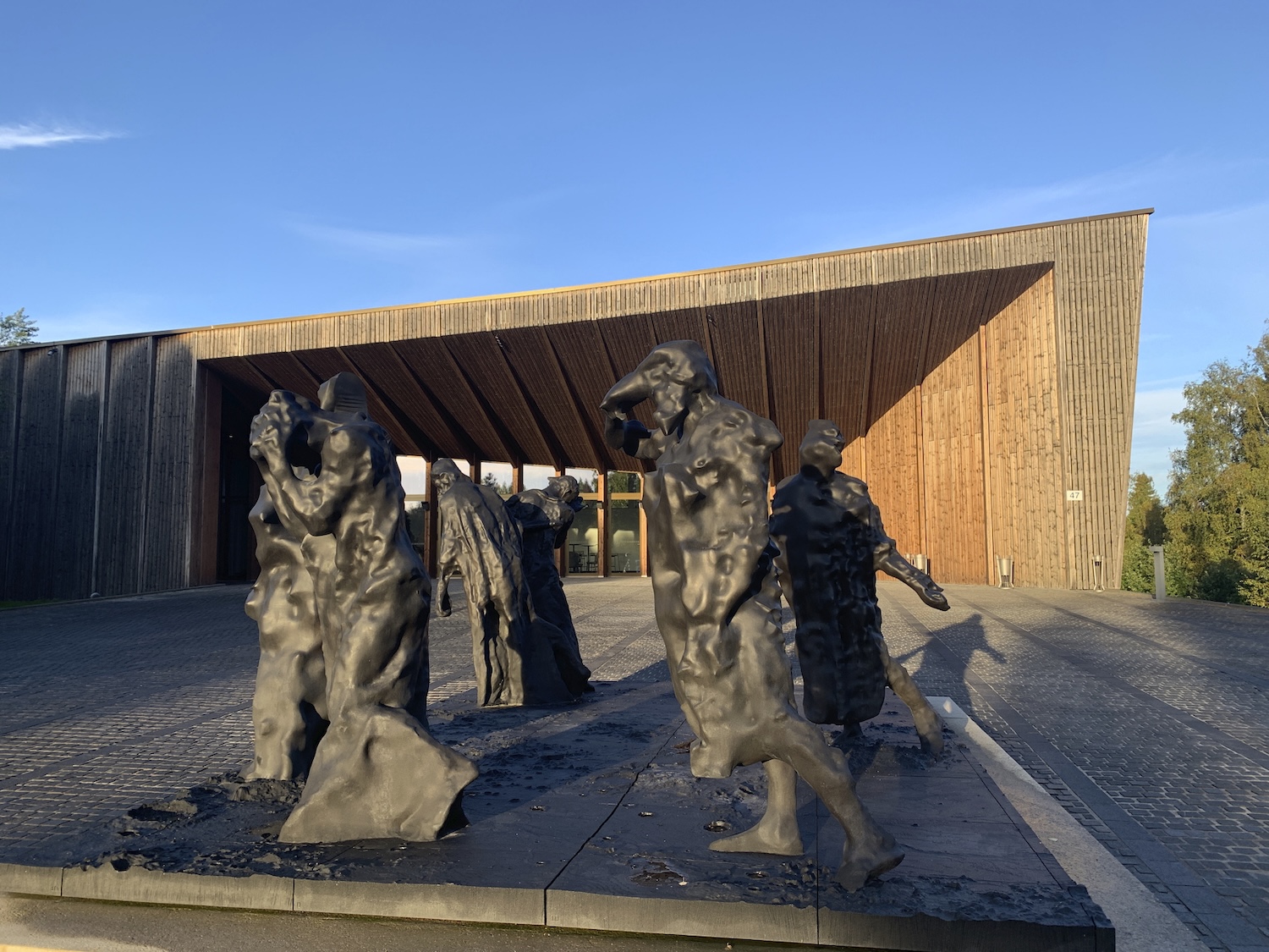 Etualalla useita ihmishahmoja esittävä veistos, taustalla näkyy puurakenteinen museorakennus, jossa on vino katto.