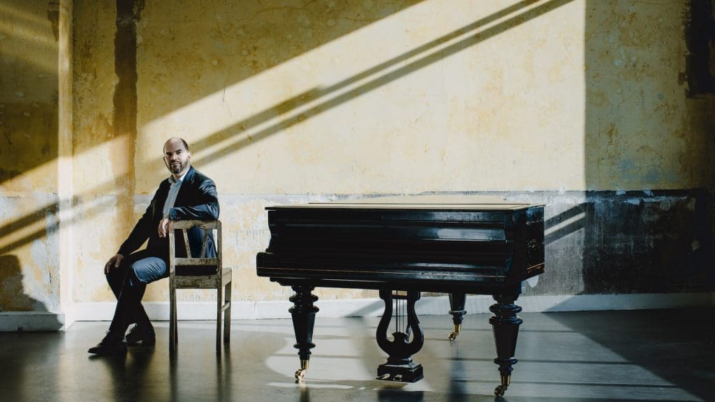 Kirill Gerstein istuu selin flyygeliin vanhalla tuolilla. Voimakas valo tulee vanhaan huoneeseen oikealta.