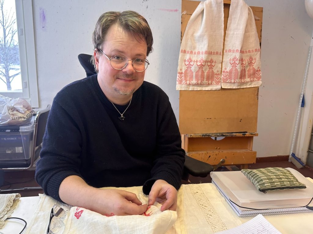 Silmälasipäinen mies tekee käsityötä pöydän ääressä ja hymyilee kameraan.