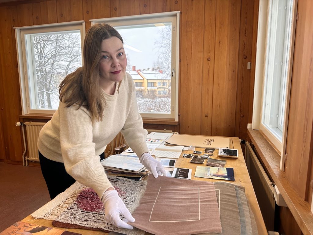 Vaaleahiuksinen nainen levittää tekstiiliä työpöydälle ja katsoo hymyillen kameraan.