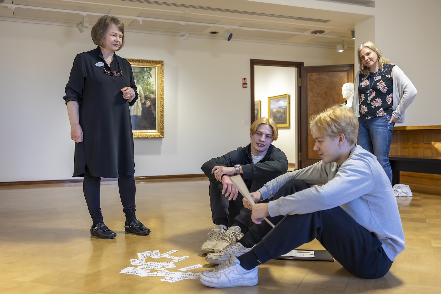 Museon opas ohjaa kahta nuorta miestä tehtävässä. Taustalla katsoo poikien opettaja.