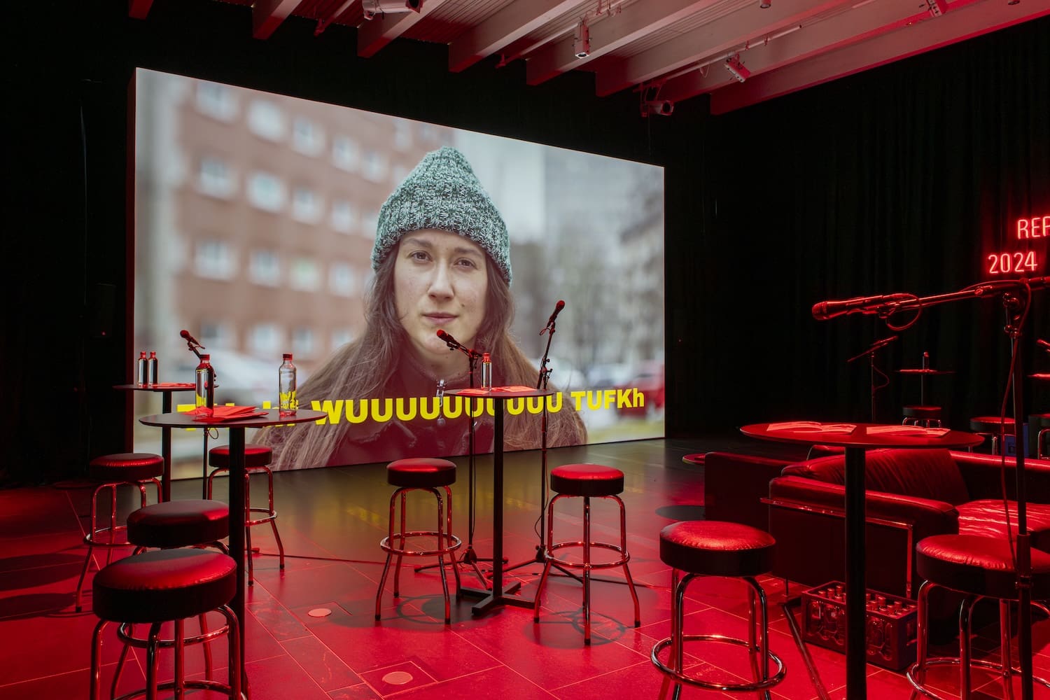 Suurella videoscreenillä näkyy nainen, joka tekee jonkinlaista sireenin ääntä.