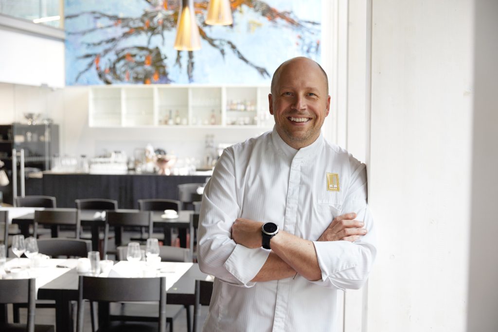 Hymyilevä mies ravintoloitsijan valkoisessa asussa nojaa seinään.