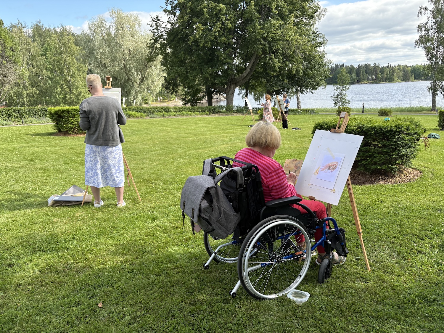 A lady sitting in a wheelchair is painting an artwork outdoors.